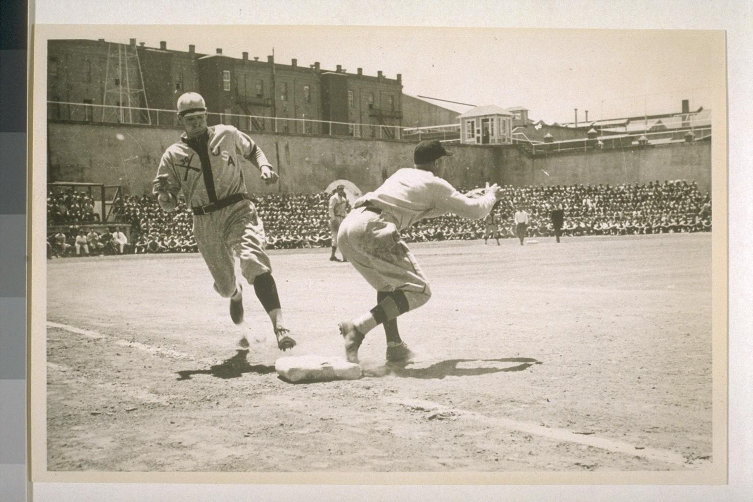Black-and-white photograph of a baseball play at the Washington State Penitentiary, runner sliding into base while inmates watch from the prison wall